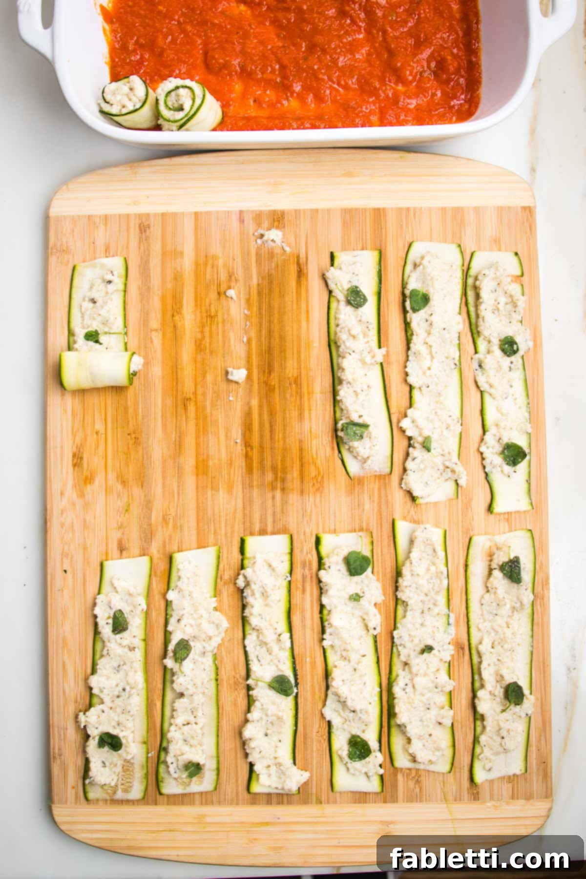 Plant-Based Zucchini Ricotta Rolls 10 Cutting board with zucchini strips spread with ricotta, one is shown to be in process of rolled up and you can see a baking pan with marinara sauce where the finished rolls are being placed.