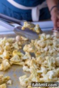 Cauliflower Florets sprinkled with spices on a baking sheet. Metal tongs are tossing the florets on the baking tray.