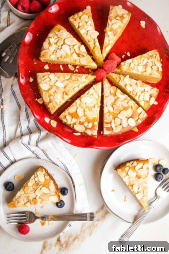 Red cake pedestal with lemon cake topped with sliced almonds. Cake is cut into pieces and there are two plates with slices of cakes plus a few berries.