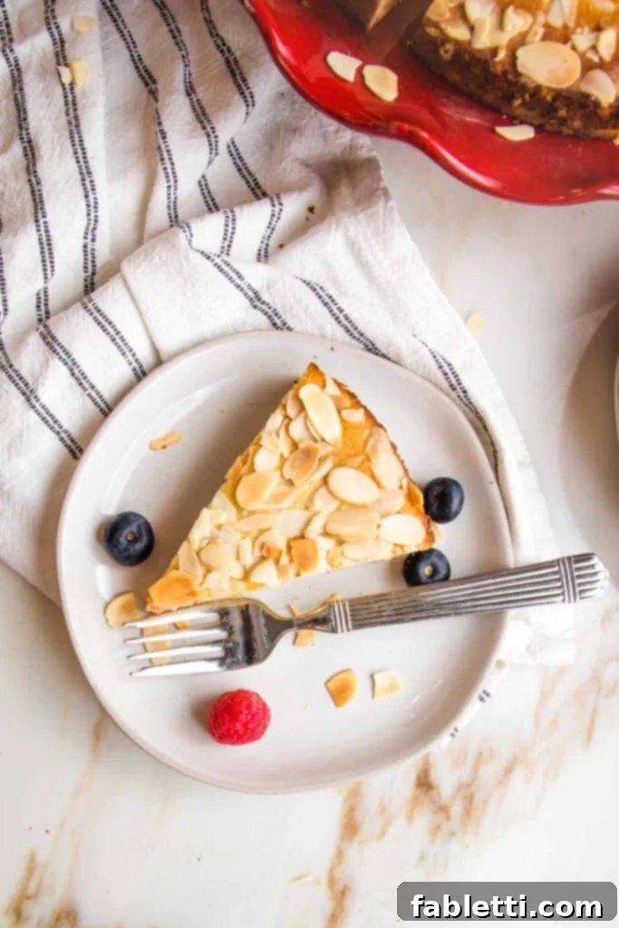 Slice of lemon cake with almonds and a few berries on a small white plate, with a fork. 