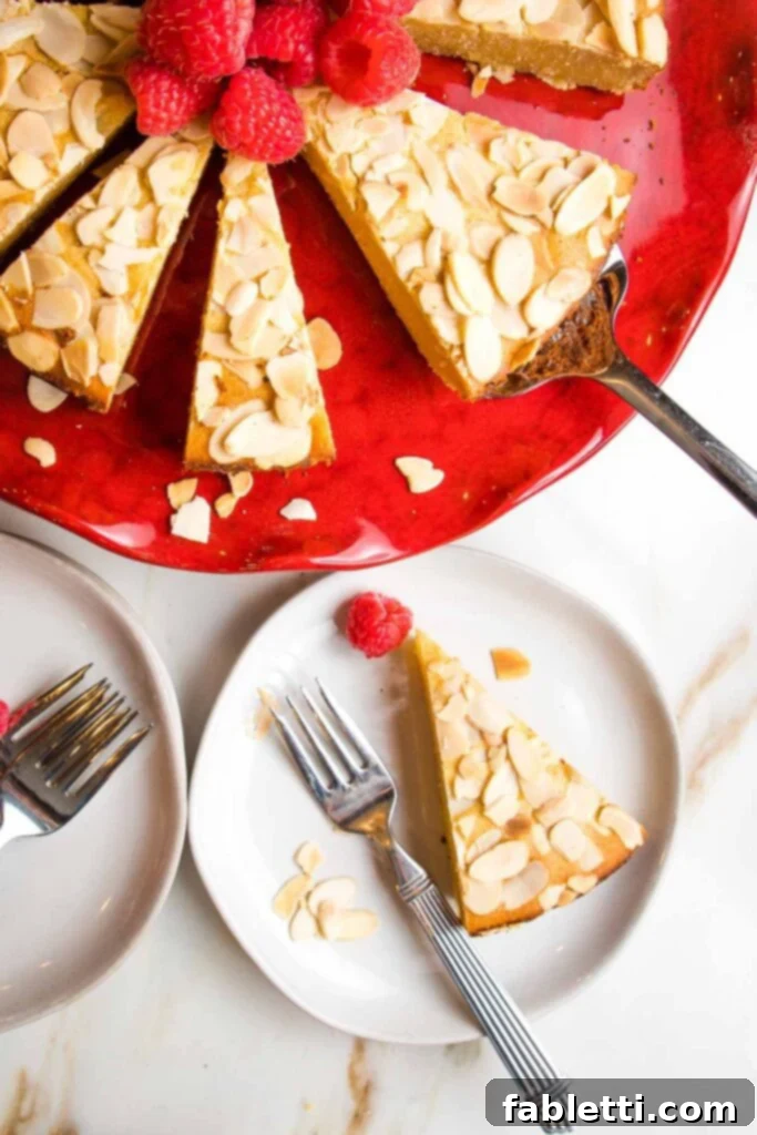 Lemon cake with sliced almonds on top of a red cake plate. There is a slice of cake on a white plate with a red raspberry. 