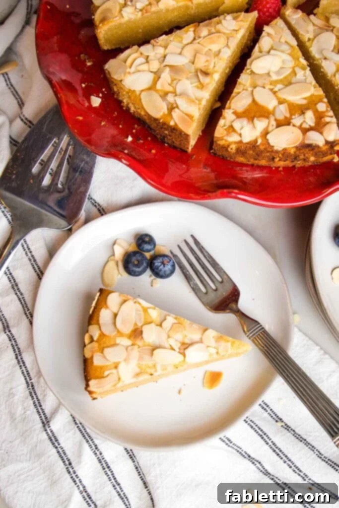 White rimmed small plate with a piece of almond lemon cake, with a few blueberries on the side. A red cake stand with remaining cake slices is on the top of the frame.