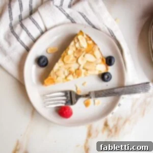 small white plate with slice of lemon cake topped with sliced almonds. Plate has a few blueberries, a raspberry and some almond slices.