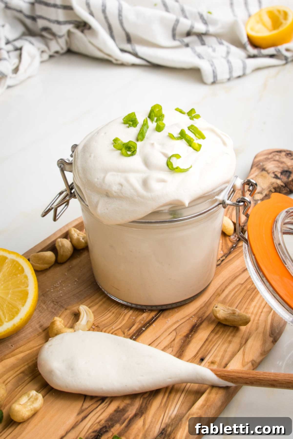 Vegan sour cream overflowing in a glass container. Highly stylized wooden spoon, lemon half and cahews.