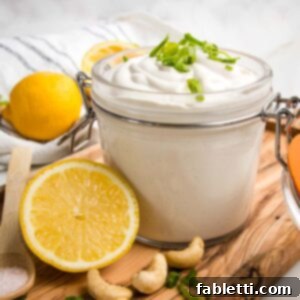 Hinged glass container filled with vegan sour cream and topped with scallions. A lemon half, some cashews and a spoon of salt on the wooden board too.