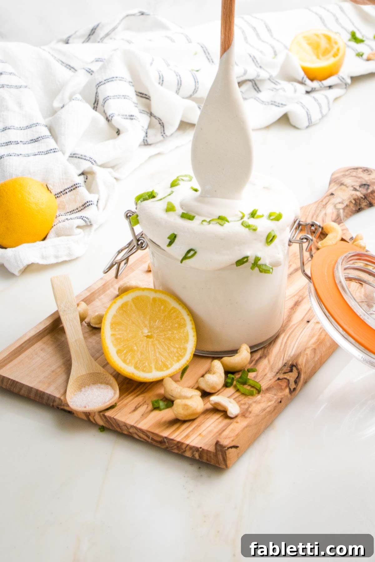 Sour cream in a glass canister. Wooden spoon dangles above canister to show creaminess of sour cream with scallions. A spoon of salt, half a lemon and raw cashews complete the picture with a dish towel in the background.