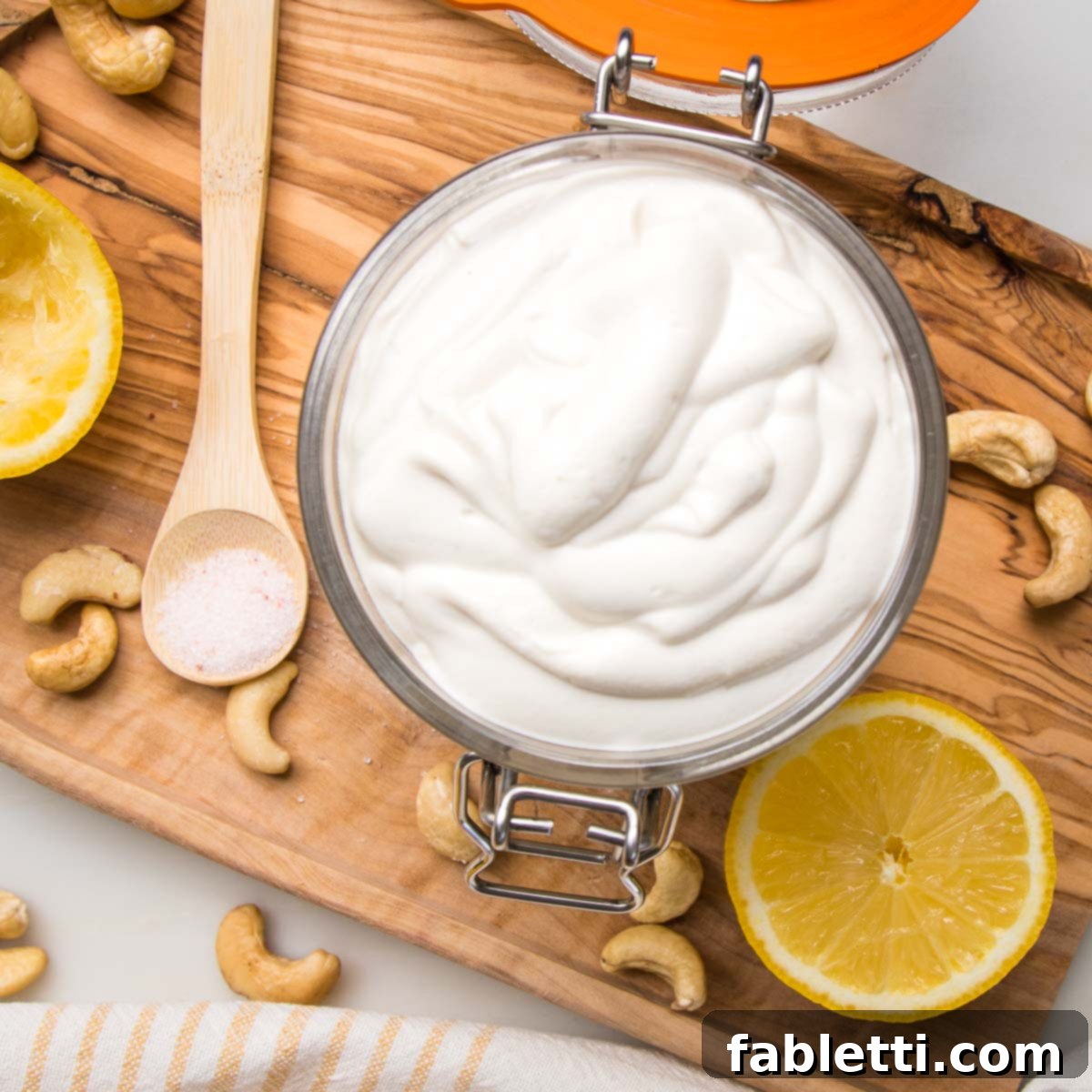 Creamy vegan sour cream in a glass container, on a wooden board. Also shows is a lemon half and one half already squeezed, a spoonful of salt and some cashews.