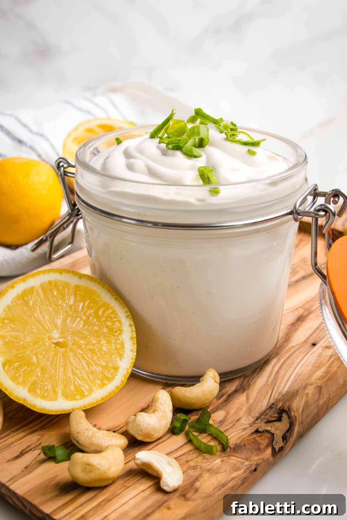 Vegan sour cream loaded into a glass canister and topped with scallions. All is on a wooden board with fresh lemon half and raw cashews. 
