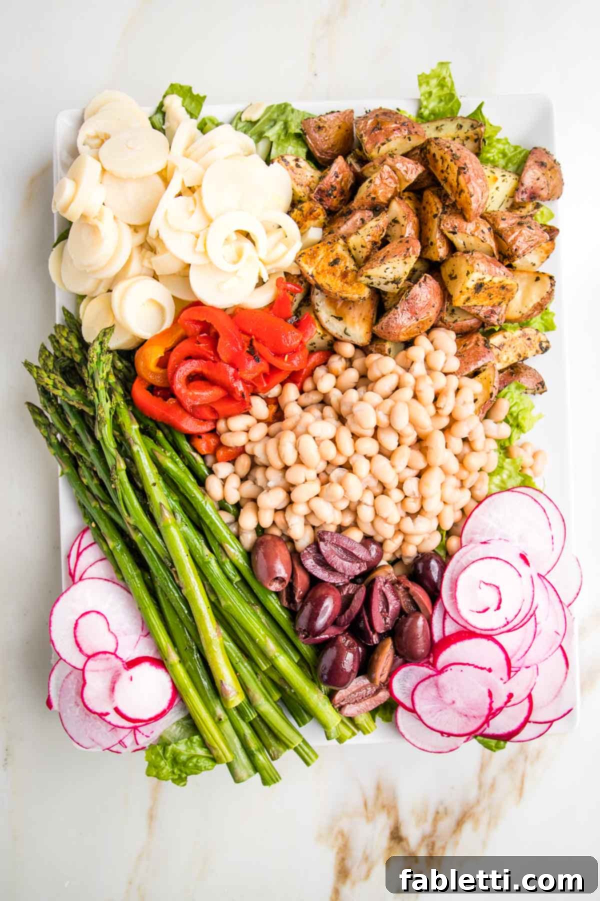 Vibrant Vegan Nicoise 11 Vegan Nicoise Salad in sections: sliced hearts of palm, herby roasted potatoes, roasted red pepper strips, asparagus, small white beans, olives and radishes on a bed of greens.