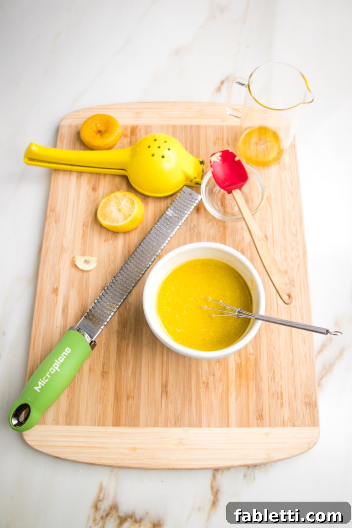 Vibrant Vegan Nicoise 10 Lemon Vinaigrette made in a small bowl with a whisk. Also on the cutting board is a lemon squeezer, zester, lemon halves, spent garlic clove and pitcher of oil.