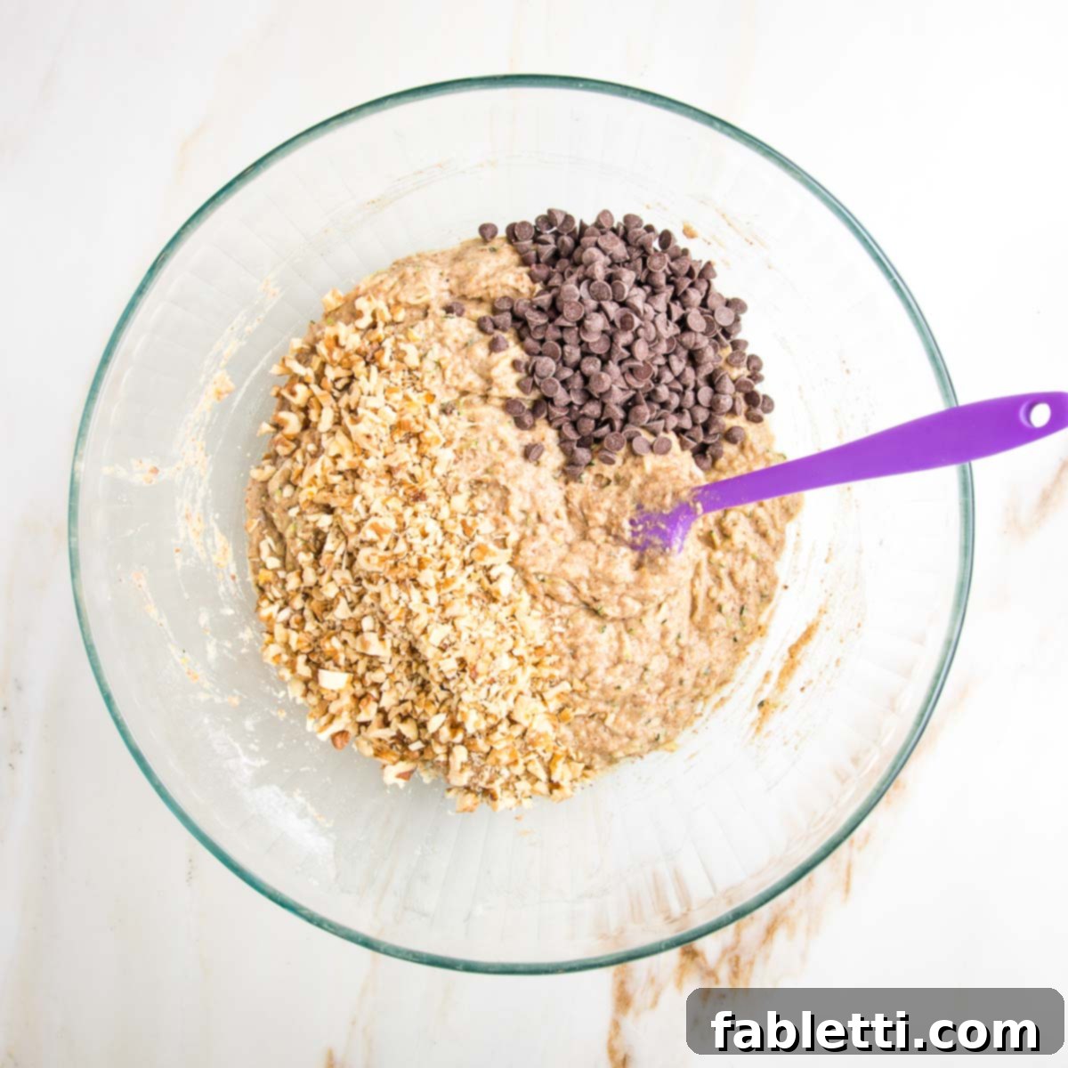 Garden Fresh Vegan Zucchini Muffins 11 Zucchini muffin batter in a glass mixing bowl with a purple spatula and mini chocolate chips plus walnuts on top.