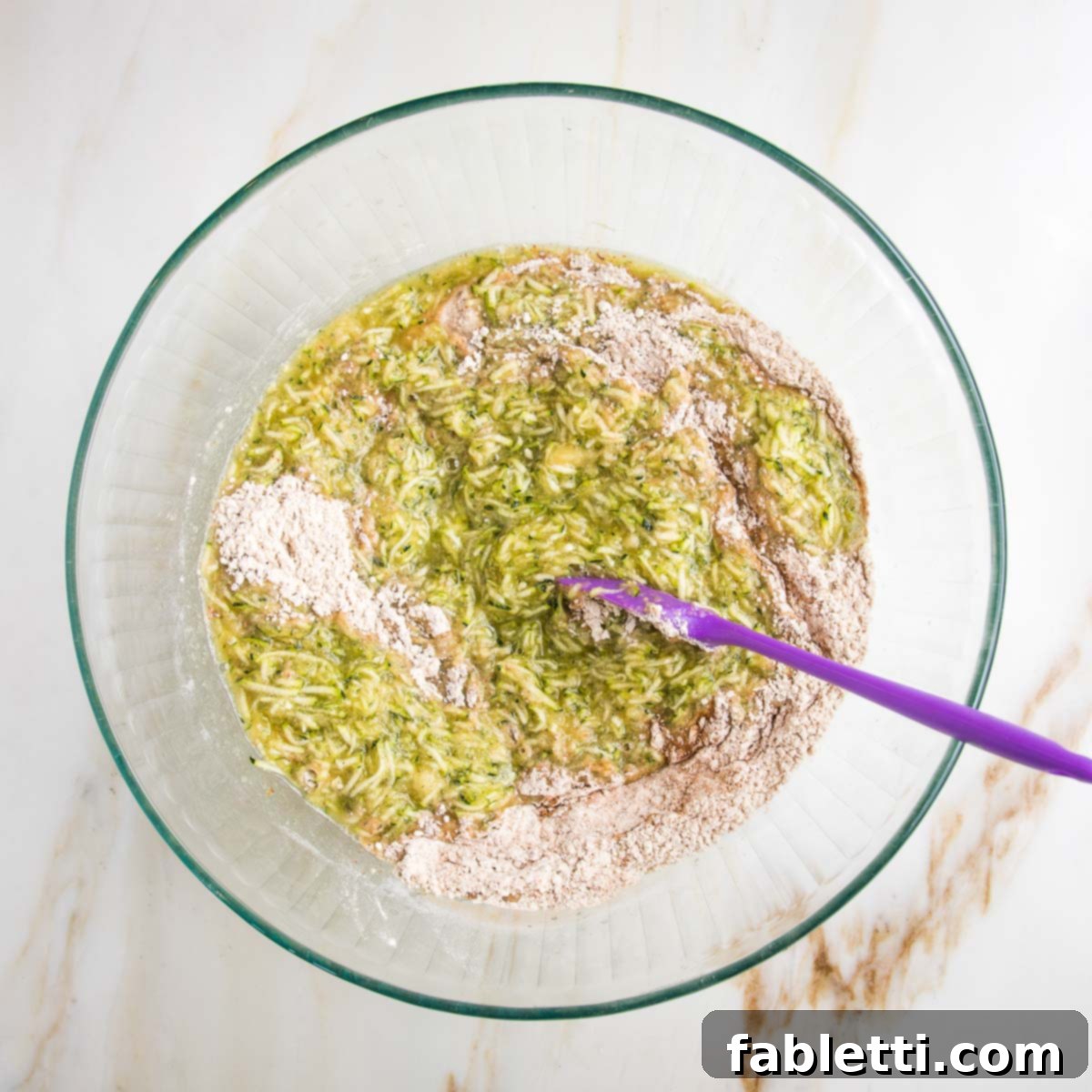 Garden Fresh Vegan Zucchini Muffins 9 Large glass mixing bowl with shredded zucchini mixture and a purple spatula mixing that together with the dry ingredients.