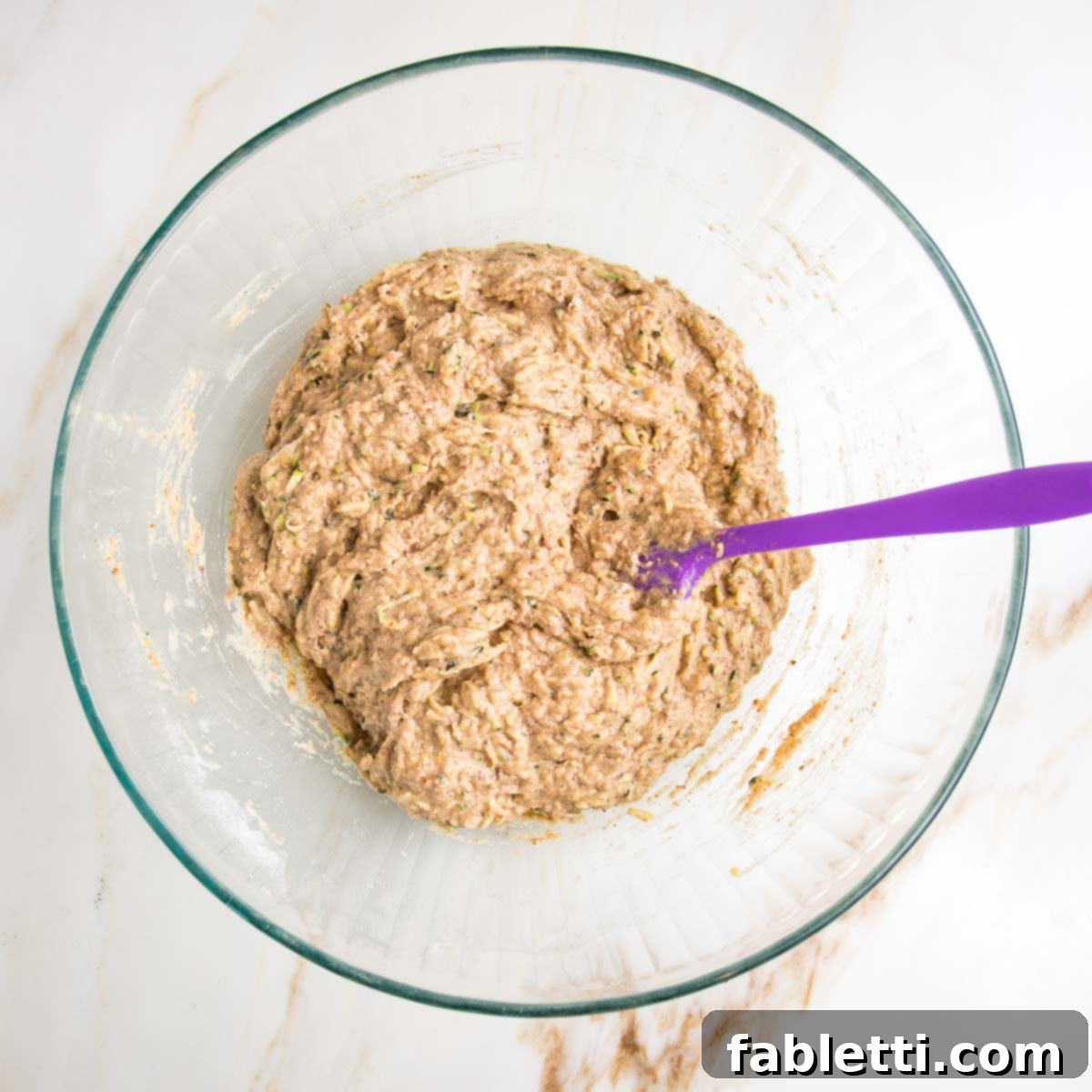 Garden Fresh Vegan Zucchini Muffins 10 Large glass mixing bowl with lumpy zucchini muffin batter and a purple spatula.