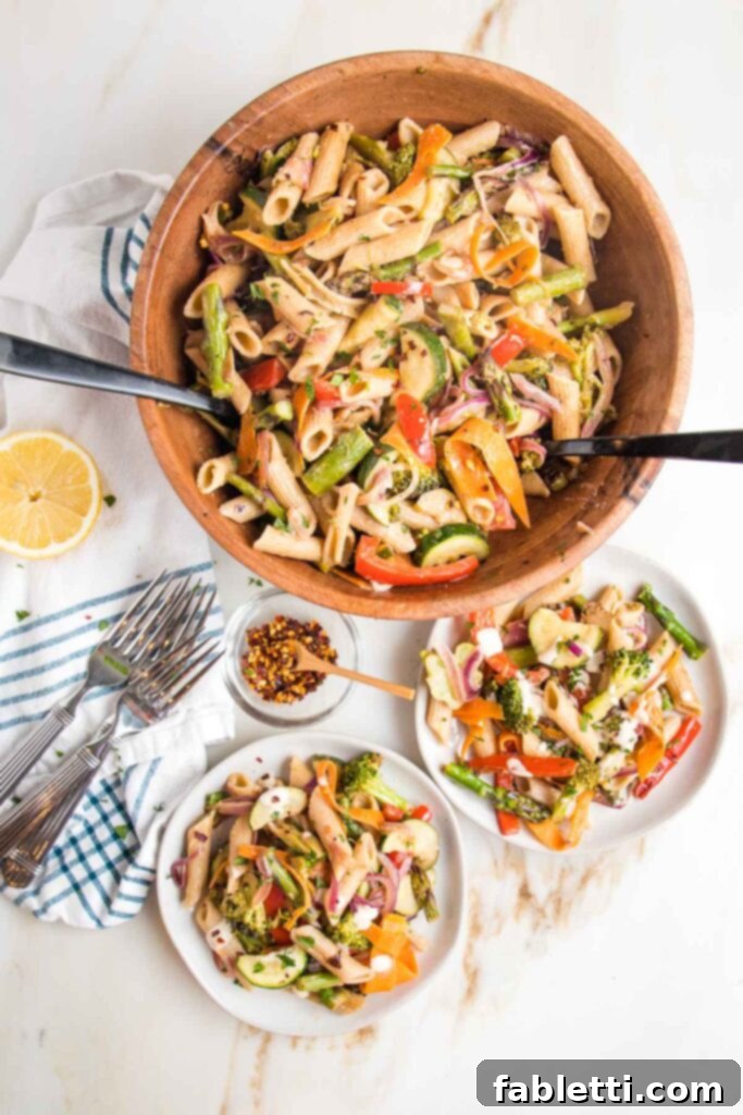 Vibrant Vegan Primavera Pasta 2 Brightly colored veggies plus penne pasta in a large wooden bowl with tossers. There are two small plates served with same vegan pasta primavera, some forks, crushed red pepper and a half a lemon.