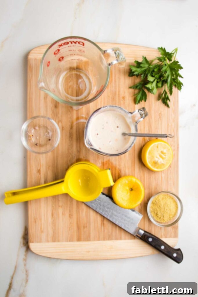 Vibrant Vegan Primavera Pasta 6 Wooden board with various glass dishes, a lemon squeezer, sharp knife, fresh parsley bunch, used up lemon halves, nutritional yeast and a pyrex measuring cup with a whisk, making a white sauce.