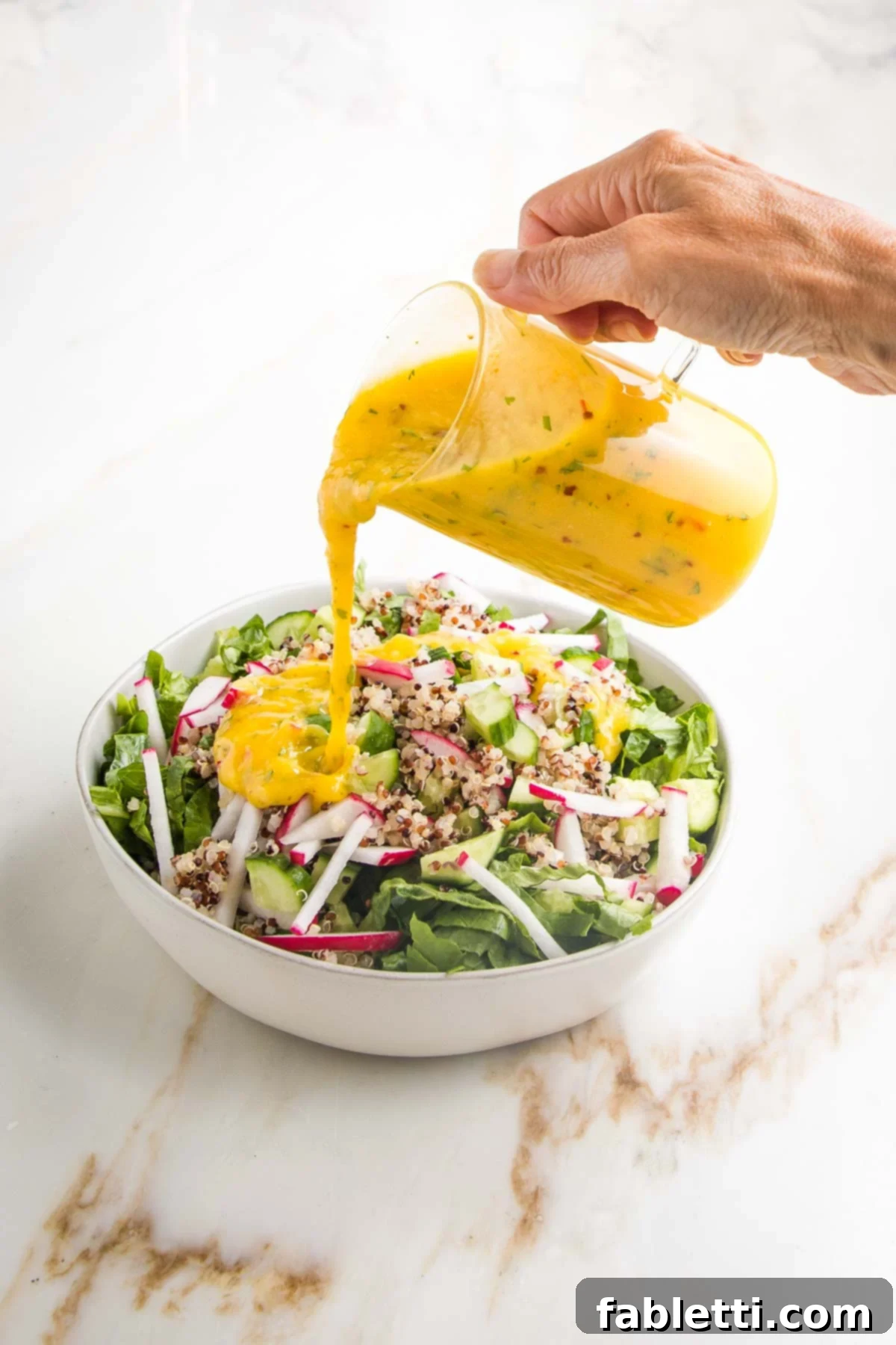Tropical Mango Vinaigrette 16 Bright yellow mango salad dressing being poured from a glass pitcher onto a green salad with radish, cucumber and quinoa.