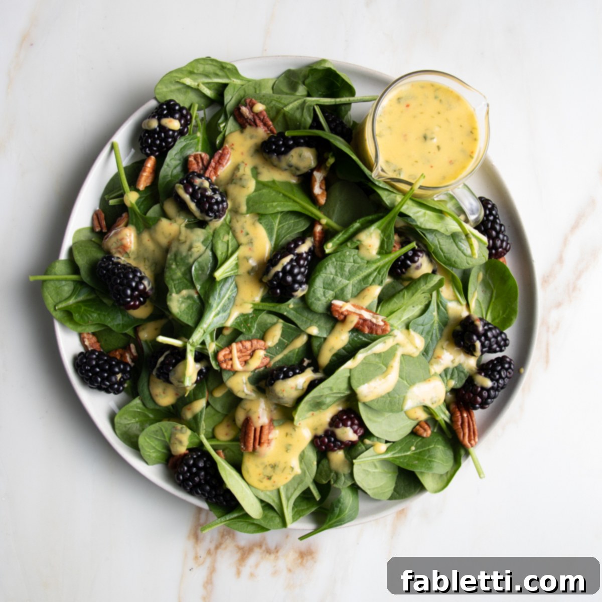 Tropical Mango Vinaigrette 17 Spinach salad with blackberries, pecans and drizzled with mango dressing. There is a small glass pitcher with extra yellow, mango dressing on the side of the round white rimmed plate.