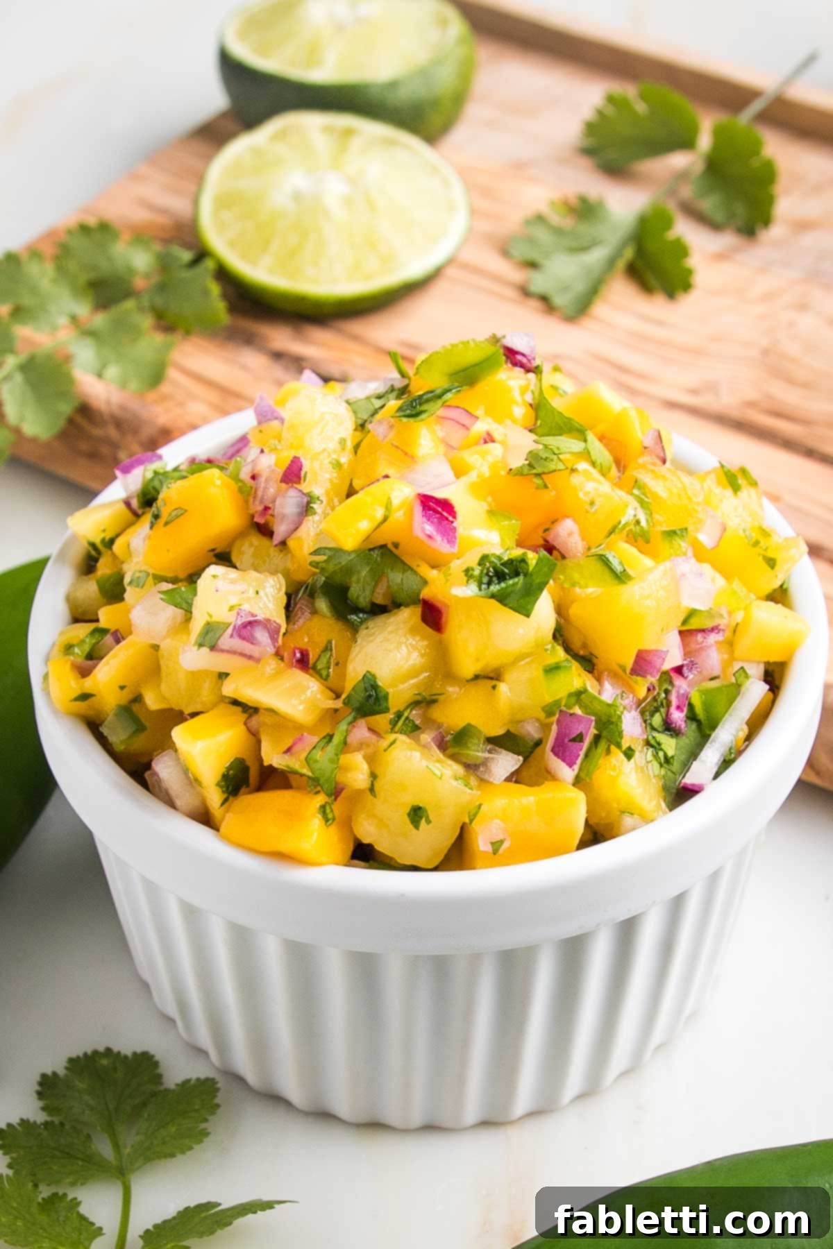 Small white bowl with pineapple mango salsa. Small pieces of bright yellow fruit, with diced red onion, jalapeno and cilantro. A juiced lime in the background with cilantro leaves on a cutting board.  