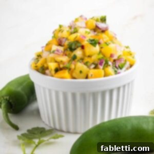 Side view of a small white bowl piled high with pineapple mango salsa. Bright yellow fruit with chunks, purple onion, green flecks of cilantro and jalapeno.