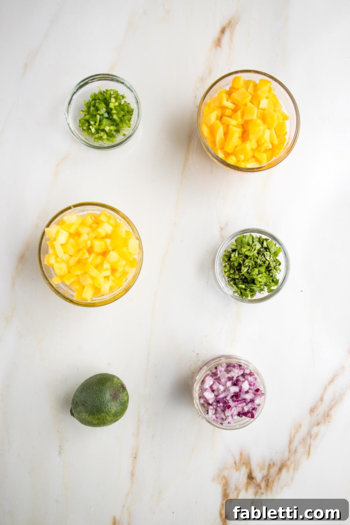 Tropical fruit salsa ingredients in small bowls: minced jalapenos, diced mango and pineapple, chopped cilantro, a lime and small diced red onions. 