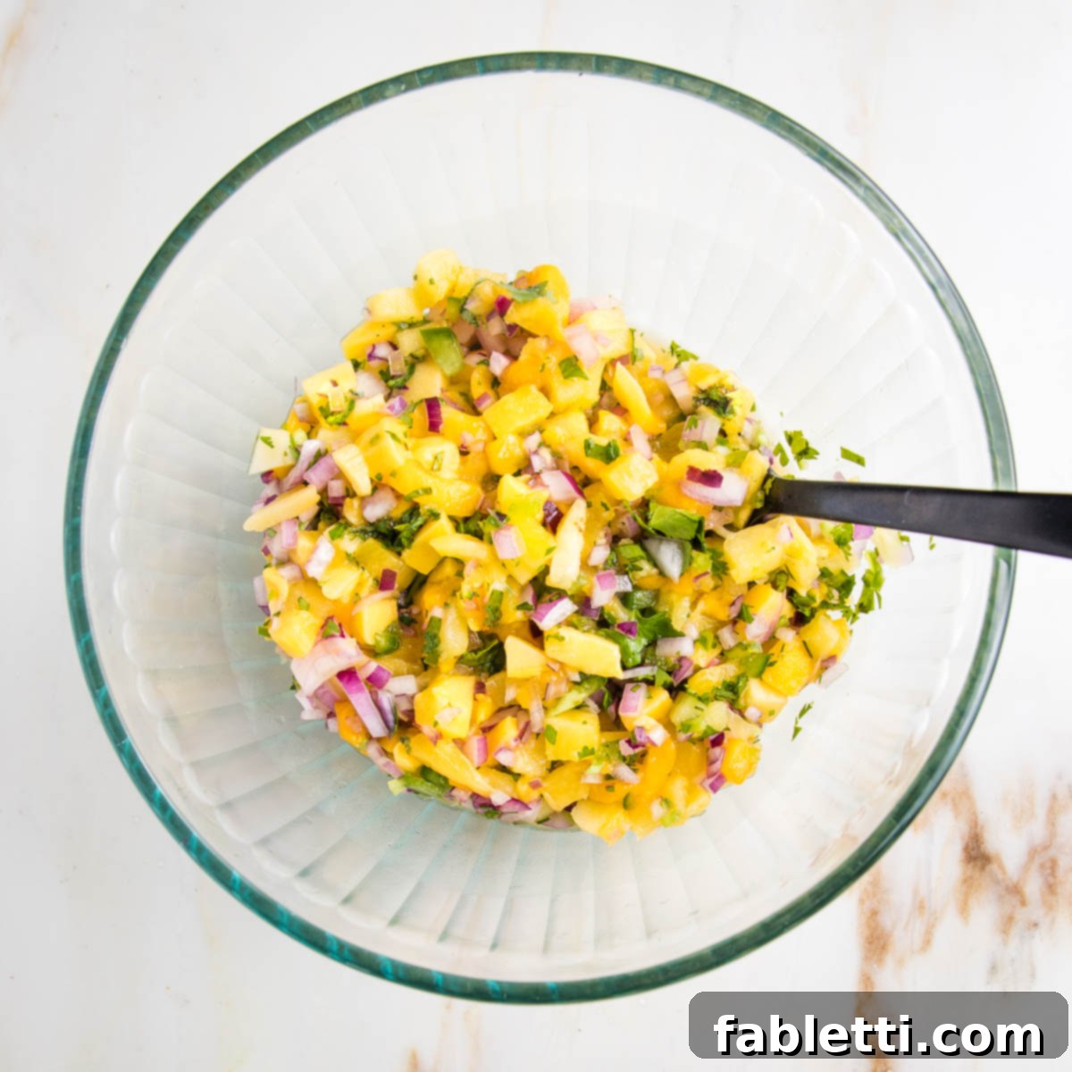 Glass bowl with colorful fruit salsa and a large spoon.  Salsa is yellow from mango and pineapple, purple onions, green jalapenos and green cilantro. 