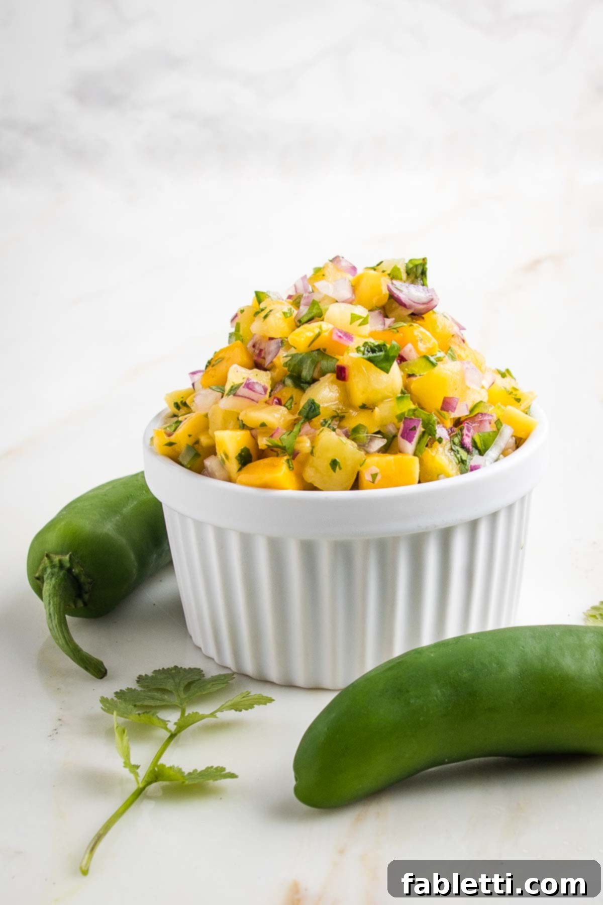 Side view of a small white bowl piled high with pineapple salsa. Jalapeno peppers and fresh cilantro in the foreground.
