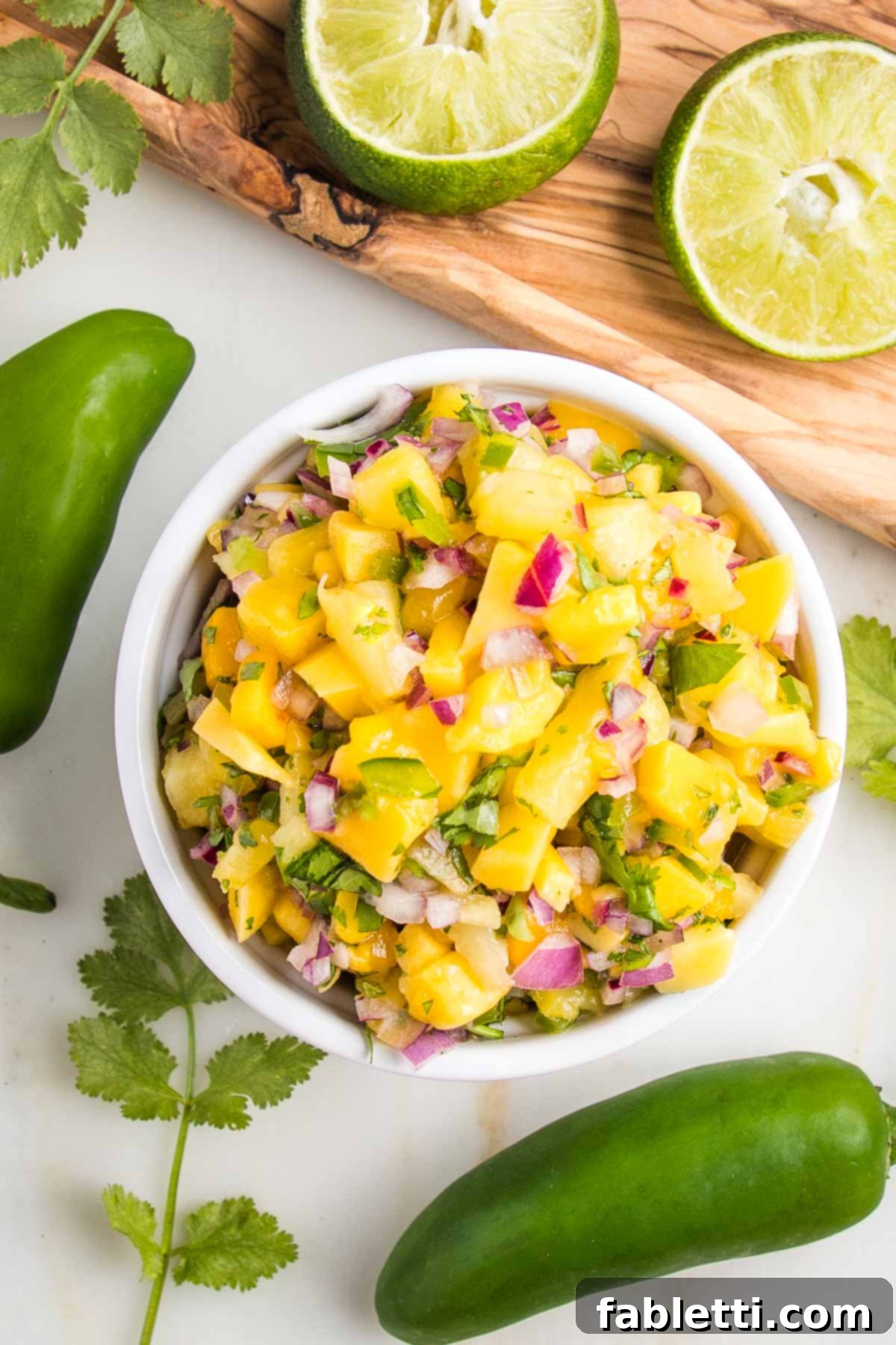 Small white bowl filled with pineapple mango salsa, whole jalapenos, sprigs of fresh cilantro and juiced limes surround the salsa.
