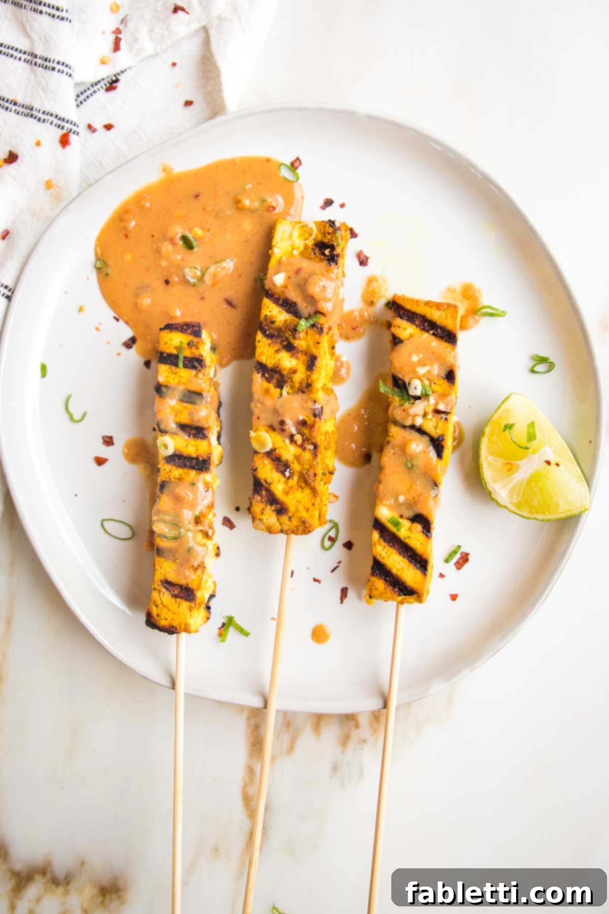 Grilled Tofu Satay with Creamy Peanut Sauce 2 Large white plate with three tofu satay skewers. Tofu has grill marks and peanut sauce is drizzled onto them. There is a lime wedge and a puddle of additional sauce.