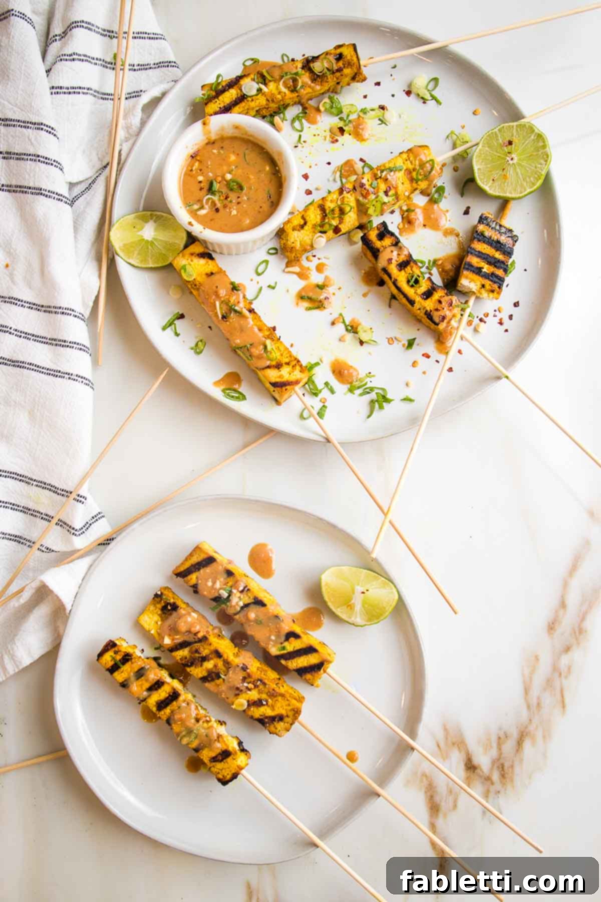 Grilled Tofu Satay with Creamy Peanut Sauce 15 Plate filled with tofu satay skewers, peanut sauce and lime wedges. Everything is sprinkled with chili flakes, sliced scallions and crushed peanuts. A smaller plate with three skewers and a lime wedge is on the bottom.