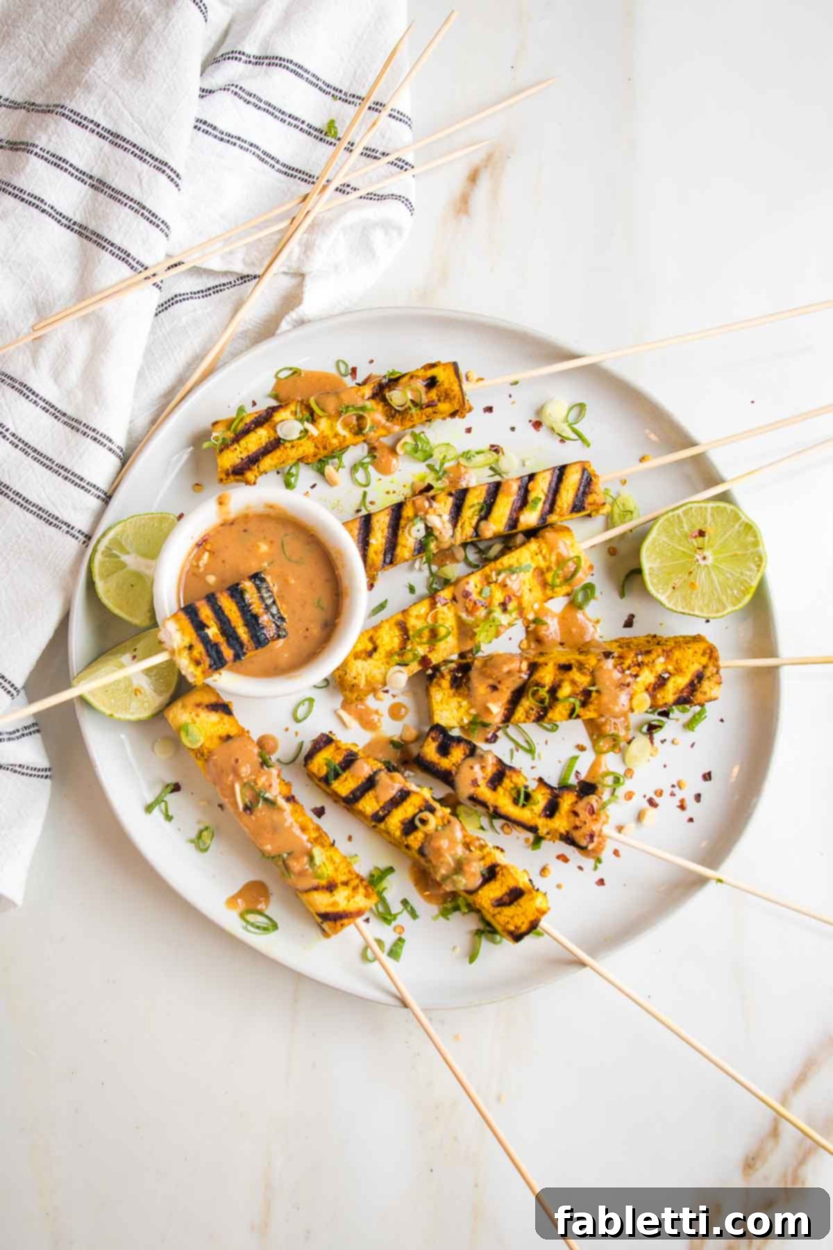 Grilled Tofu Satay with Creamy Peanut Sauce 16 Plate of grilled tofu satay skewers with a small dish of peanut sauce, sauce drizzled onto skewers and everything is sprinkled with scallions, peanuts, crushed red pepper and more sauce.