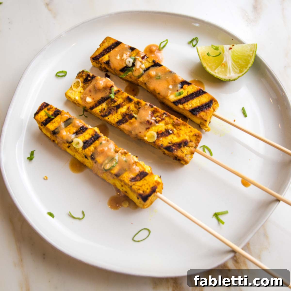 Grilled Tofu Satay with Creamy Peanut Sauce 3 Three tofu skewers with grill marks on a plate, drizzled with peanut sauce.