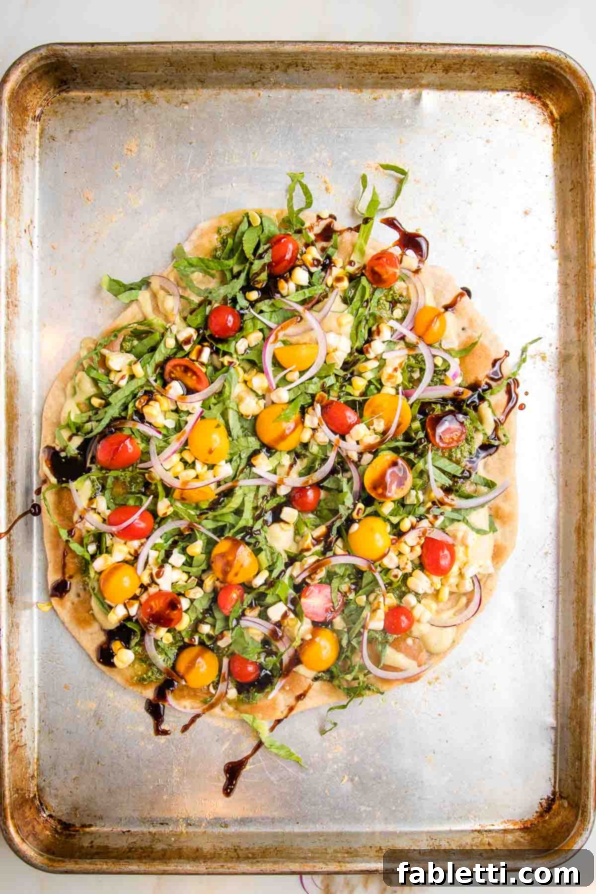 Effortless Yeast-Free Flatbread Pizza 16 A colorful flatbread pizza on a baking tray, generously topped with pesto, hummus, red and yellow tomatoes, chopped greens, and a balsamic drizzle, ready to be enjoyed.