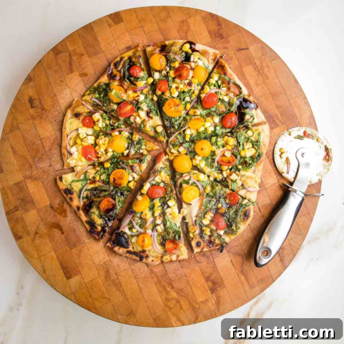 Effortless Yeast-Free Flatbread Pizza 18 A flatbread pizza, vibrantly topped with red and yellow tomatoes, fresh greens, onions, and pesto, neatly sliced on a wooden cutting board with a pizza cutter beside it.