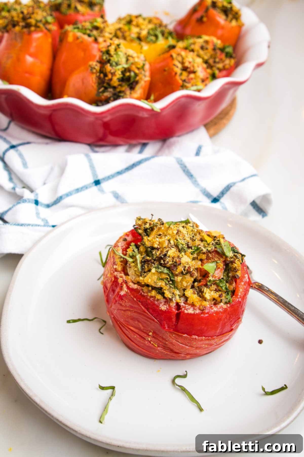 Plant-Powered Stuffed Tomatoes 2 Baked stuffed tomato, sprinkled with fresh basil. In the background is a red pie dish filled with more baked stuffed tomatoes, ready to be served.