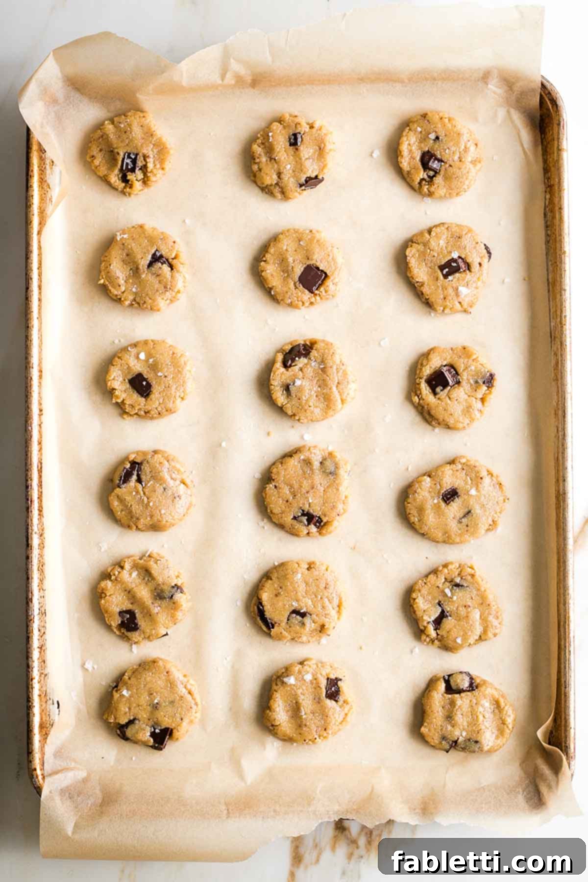 Chewy Vegan Gluten-Free Tahini Chocolate Chip Cookies 12 Rows of chocolate chunk cookies flattened on a tray and sprinkled with flakey sea salt.