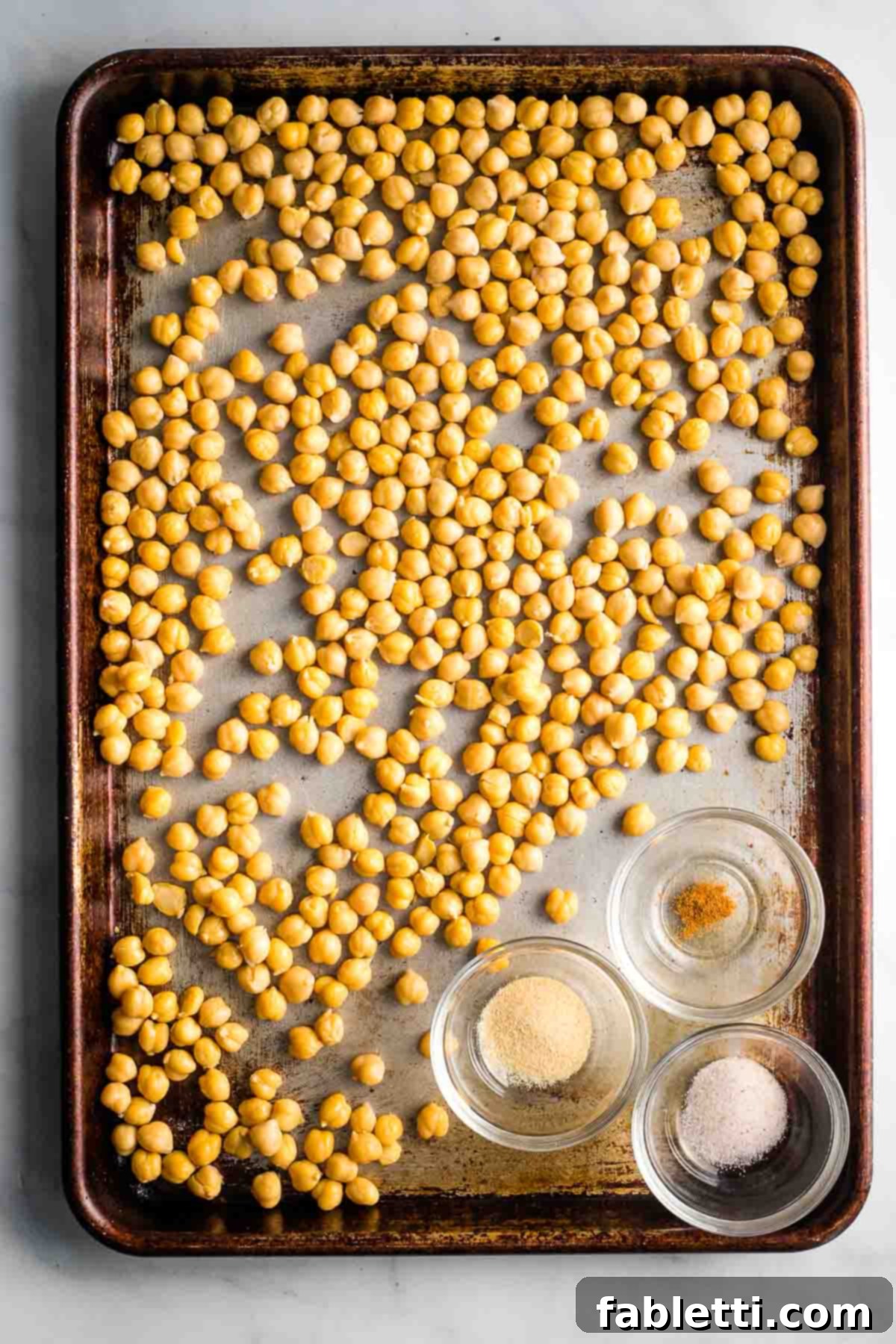 Golden Crispy Chickpea Bites 5 Chickpeas in a single layer on a baking sheet with three small dishes with spices.