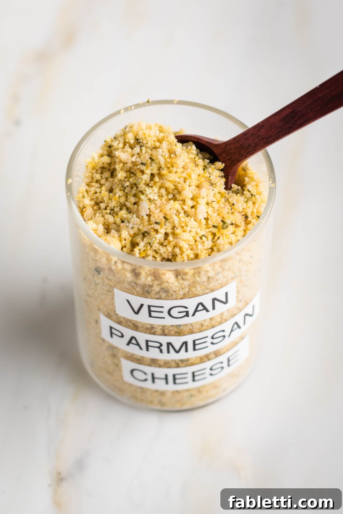Nut-Free Plant-Based Parmesan 2 Glass spice jar labeled "vegan parmesan cheese" showing the yellow mixture with grated cheese texture from the top with a spoon.