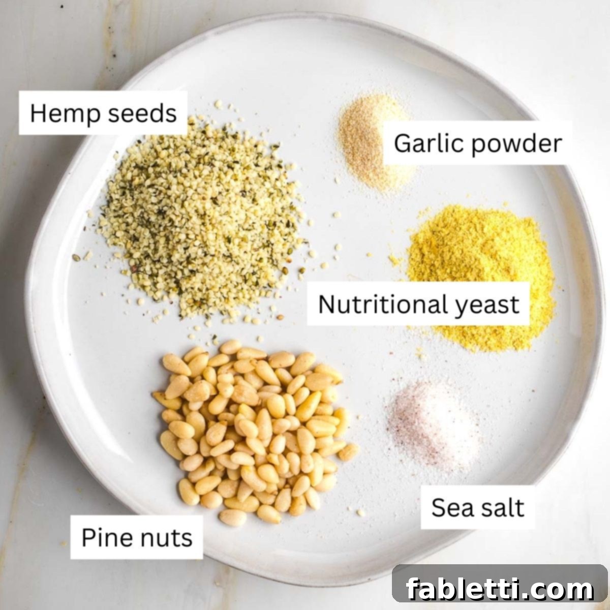 Nut-Free Plant-Based Parmesan 3 Labeled ingredients sprinkled onto a plate: pine nuts, hemp seeds, garlic powder, nutritional yeast, salt.