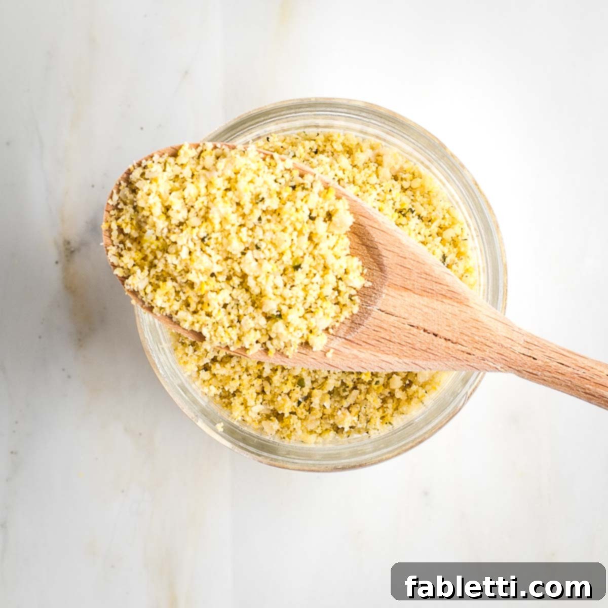 Nut-Free Plant-Based Parmesan 7 Glass jar of parmesan with a wooden spoonful of the yellow textured mix resting on top.