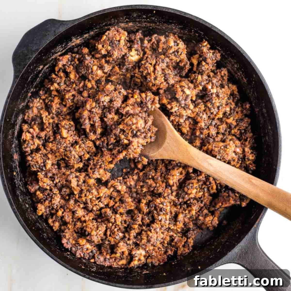 Umami-Packed Vegan Mushroom Lentil Crumble 11 The vegan ground beef simmers in a cast iron skillet, a wooden spoon resting beside it. The mixture has achieved a beautiful browned and crumbly texture, perfectly resembling traditional ground beef.