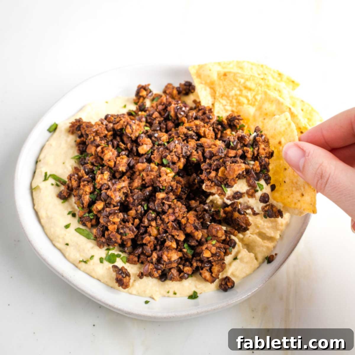Umami-Packed Vegan Mushroom Lentil Crumble 12 A tortilla chip is scooping up a generous portion of creamy hummus topped with savory vegan ground beef, highlighting a delicious and easy serving suggestion.