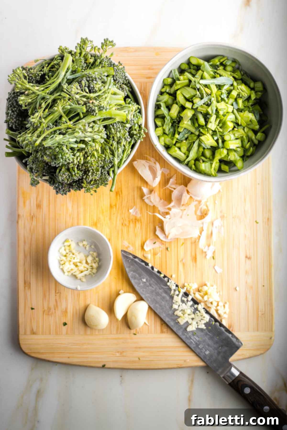 Knife mincing garlic on a board that has bowls of trimmed broccolini.