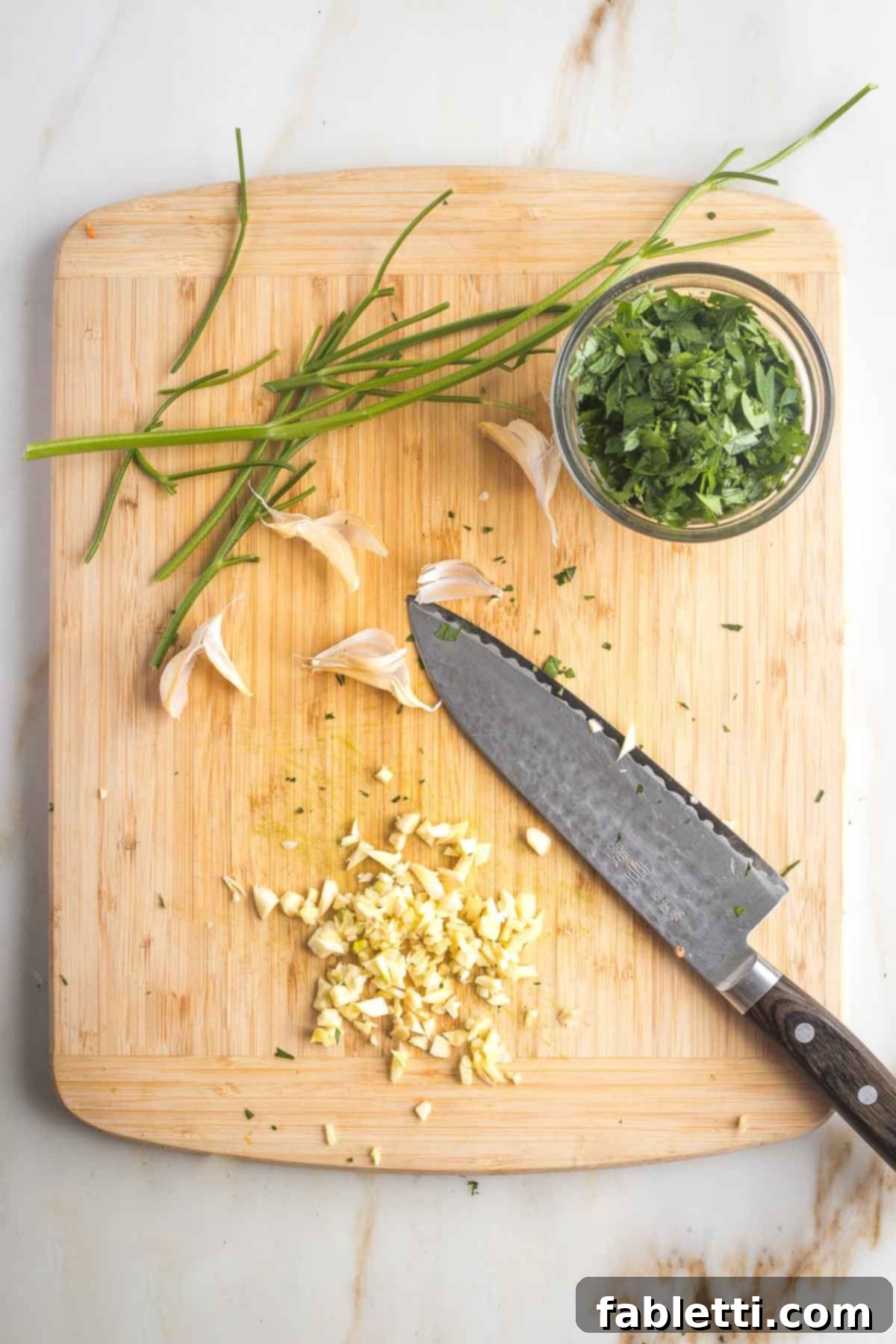 Savory Sautéed Eggplant Salad 9 A small dish of chopped parsley next to a knife roughly chopping fresh garlic cloves on a cutting board.