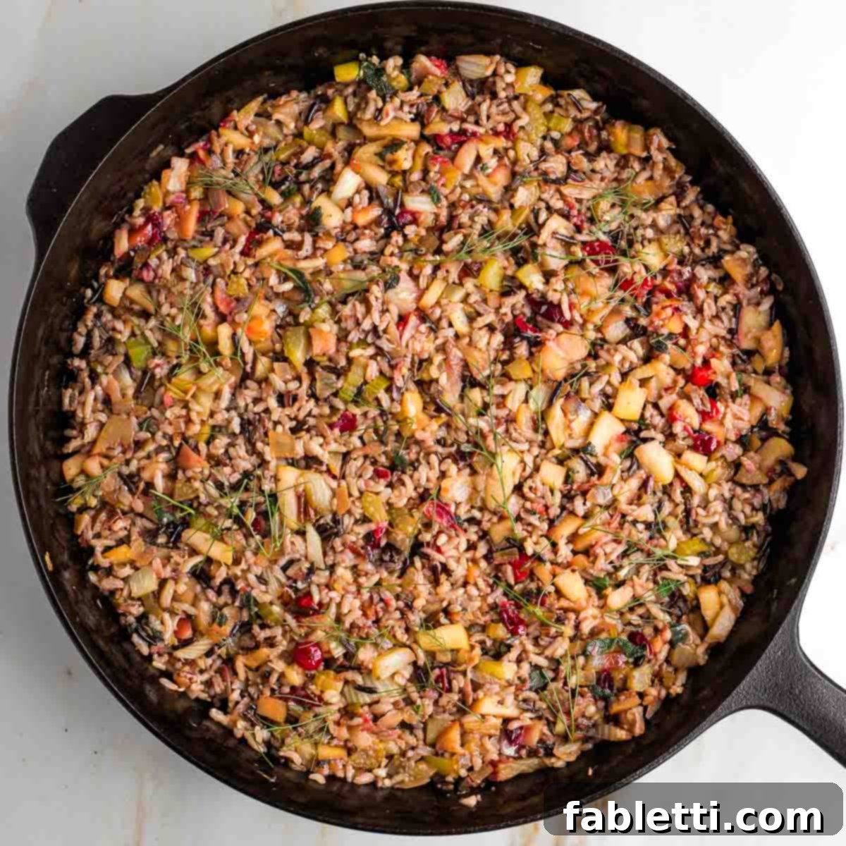 Colorful wild rice stuffing, featuring bright red cranberries and fresh green herbs, mixed together in a skillet.