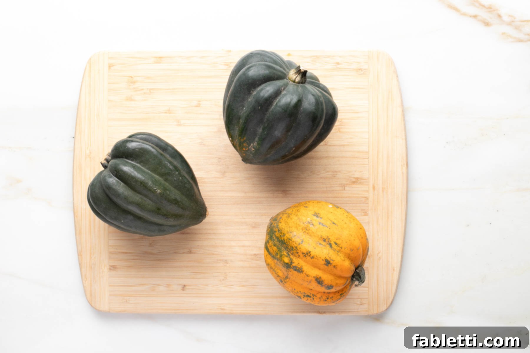 Three acorn squash arranged on a wooden cutting board; two are deep green, and one shows a lovely speckled orange and green hue, indicating optimal ripeness.