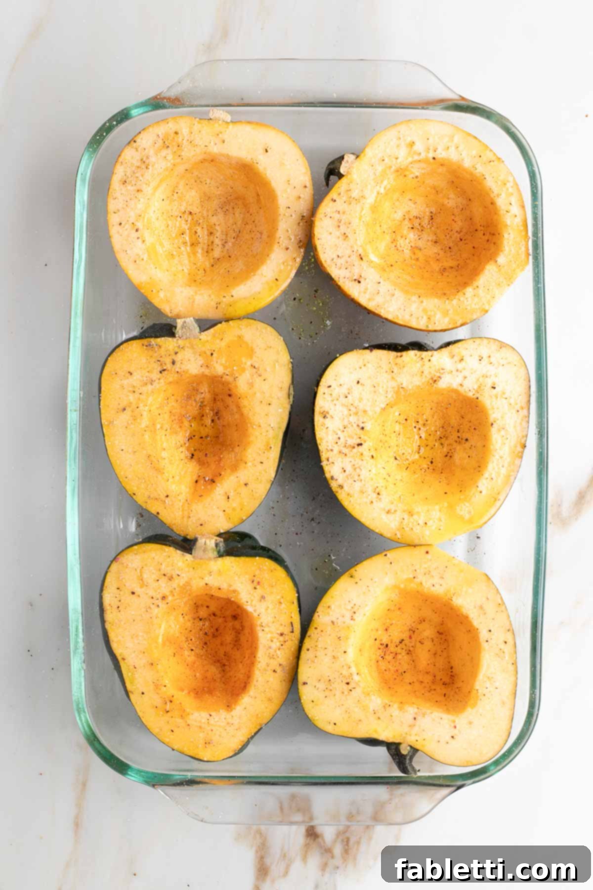 Acorn squash halves arranged cut-side up in a baking dish, lightly drizzled with olive oil and seasoned with salt and pepper.