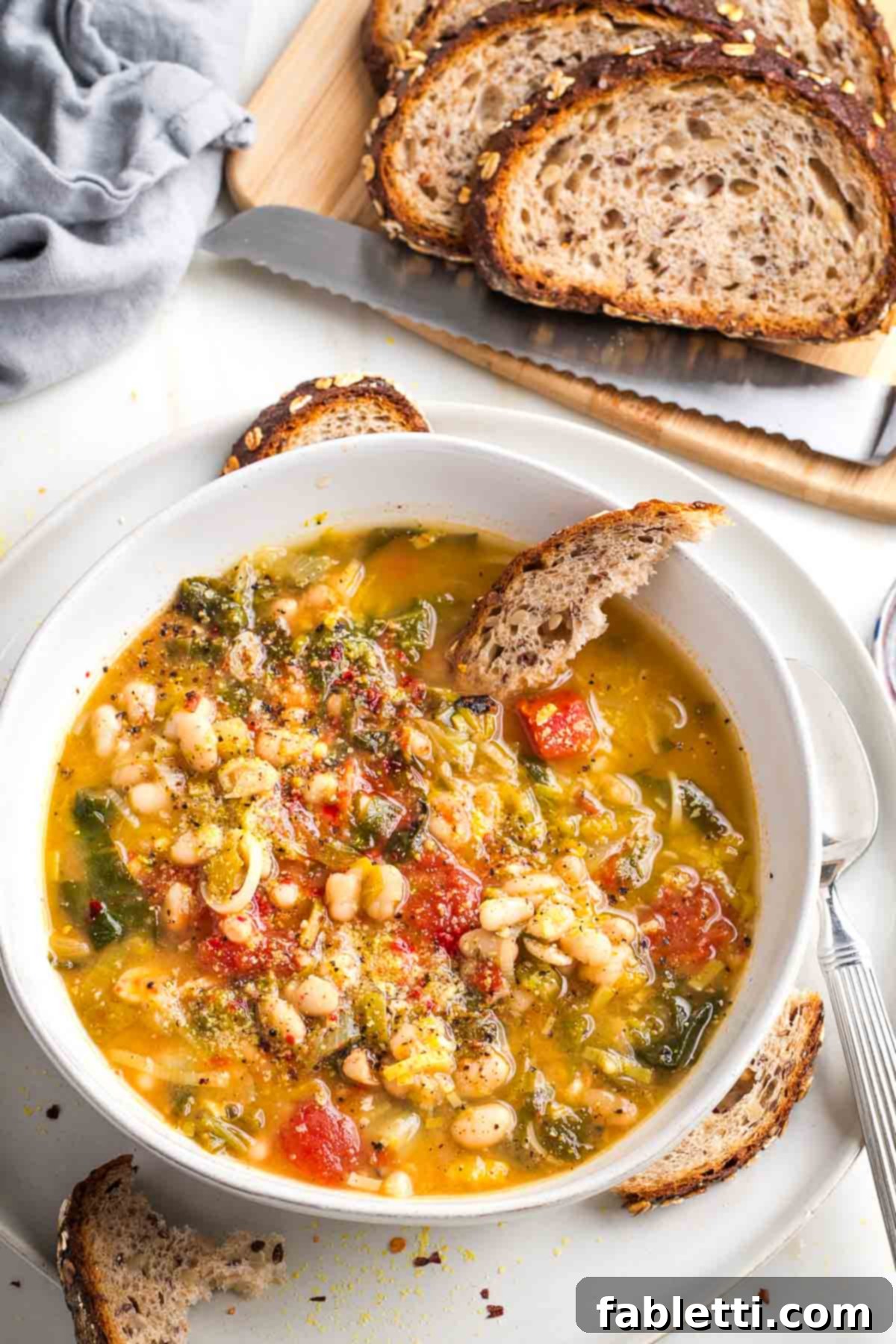 Chunky white bean, escarole and tomato soup with sliced sourdough in the background and a piece for dunking into the bowl.