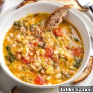 Chunky soup with tomatoes, escarole and beans in a yellow broth with a wedge of sourdough dipping into the bowl.
