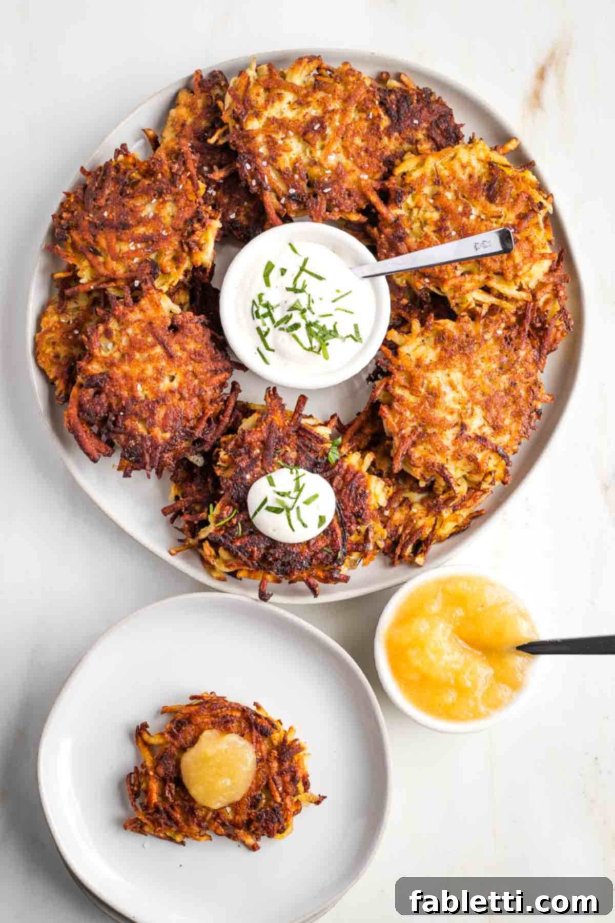 Grandma's Golden Potato Latkes 2 A festive plate of golden-brown potato latkes, with a bowl of sour cream in the center, and a side plate featuring a single latke with applesauce.