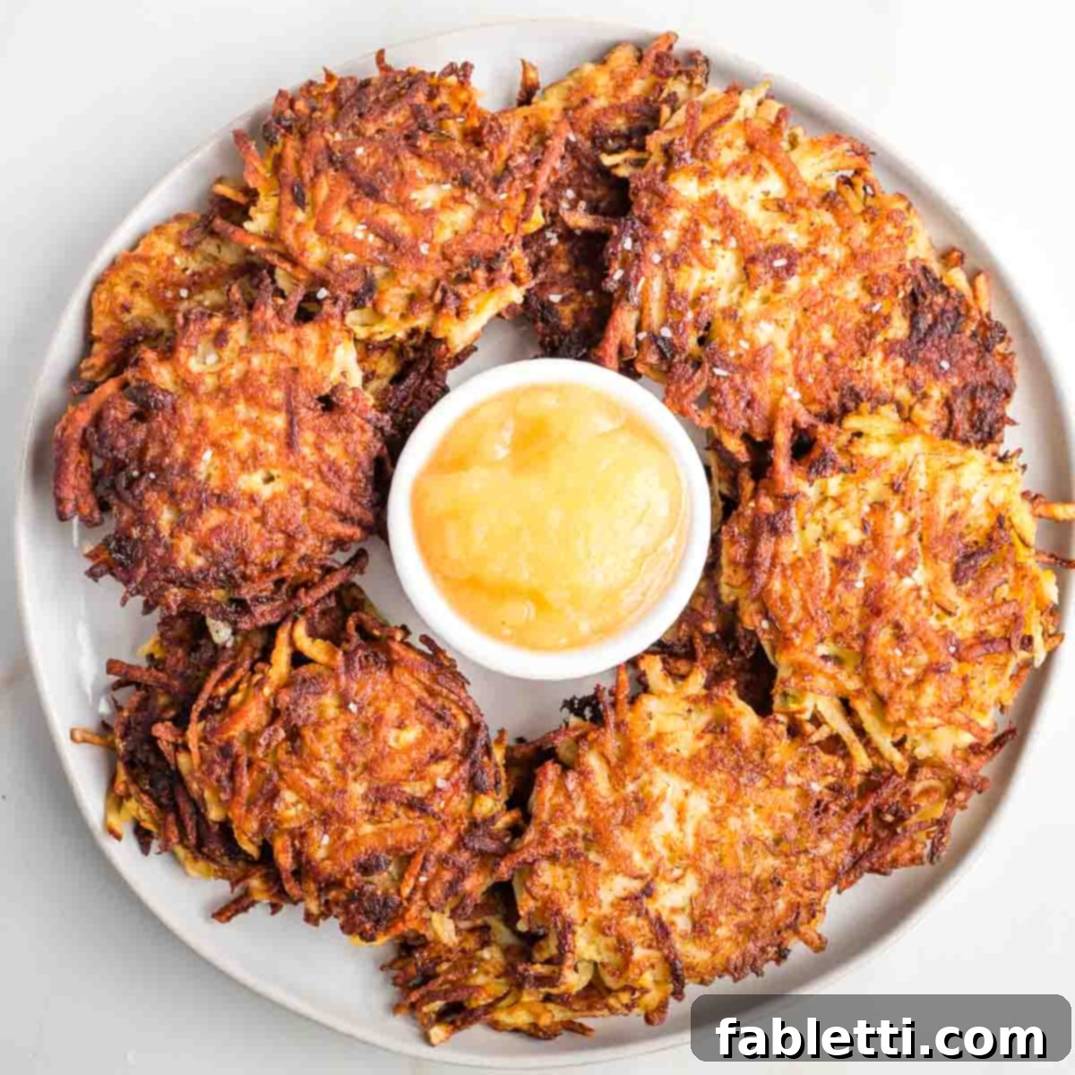 Grandma's Golden Potato Latkes 16 A beautiful platter of perfectly crispy potato latkes, with a small dish of homemade applesauce in the center, ready to be enjoyed.